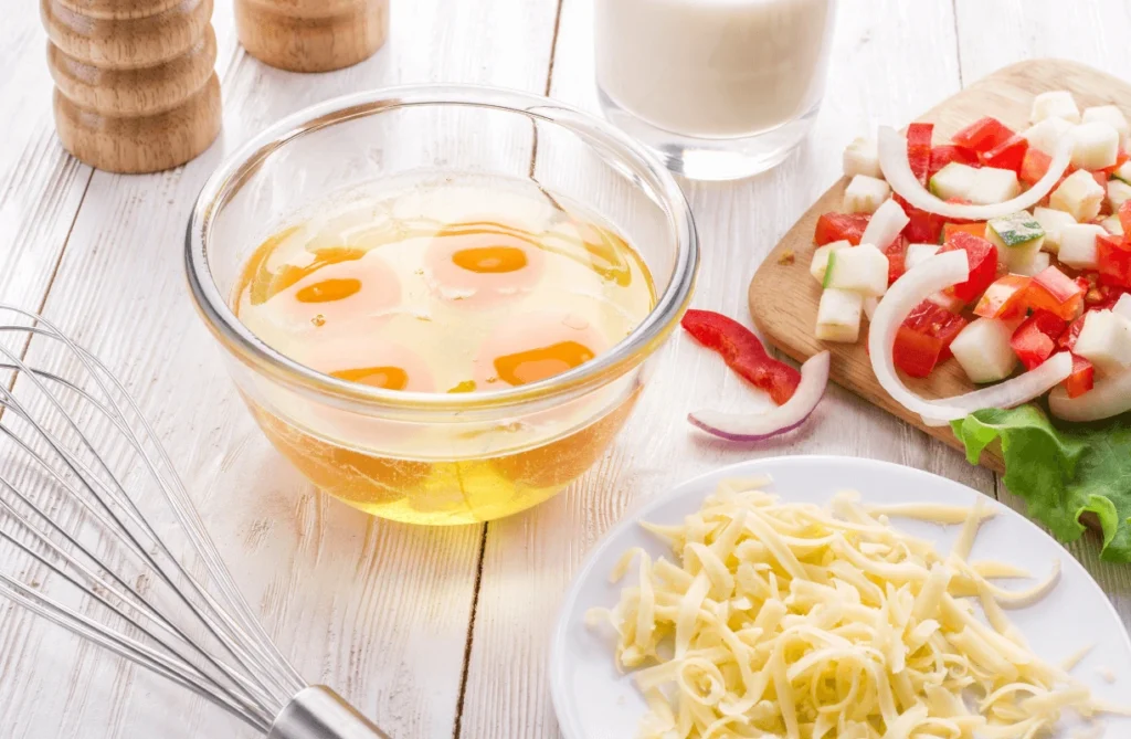 Fresh eggs, vegetables, and shredded cheese arranged on a countertop for making an omelette with cheese and vegetables.