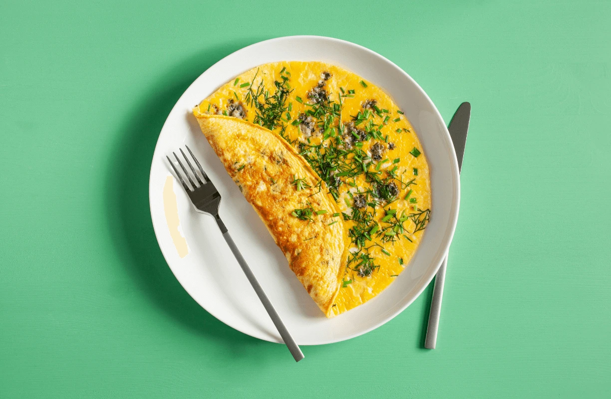 A fluffy omelette served on a white plate, topped with herbs, alongside a fork and knife on a green background.